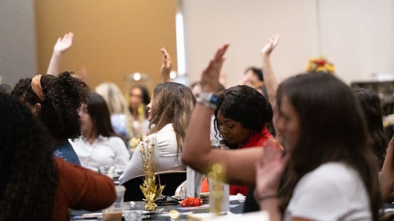 Attendees at an event raise their hands during a group activity, seated at tables with festive gold centerpieces.
