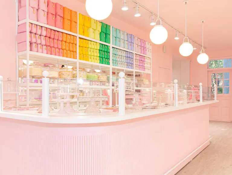 Interior of the Stoney Clover Lane retail store featuring a pastel pink counter and walls, with shelves displaying neatly arranged bags in a vibrant rainbow color gradient. The space is brightly lit with globe pendant lights and has a whimsical, candy-shop aesthetic.