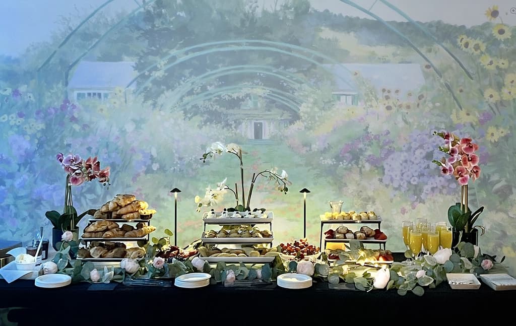 A beautifully arranged breakfast buffet with pastries, fruits, and juice set up against a scenic floral backdrop, adorned with orchids and flowers.