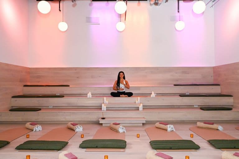Woman meditating in a serene studio with mats, bolsters, candles, and soft ambient lighting.