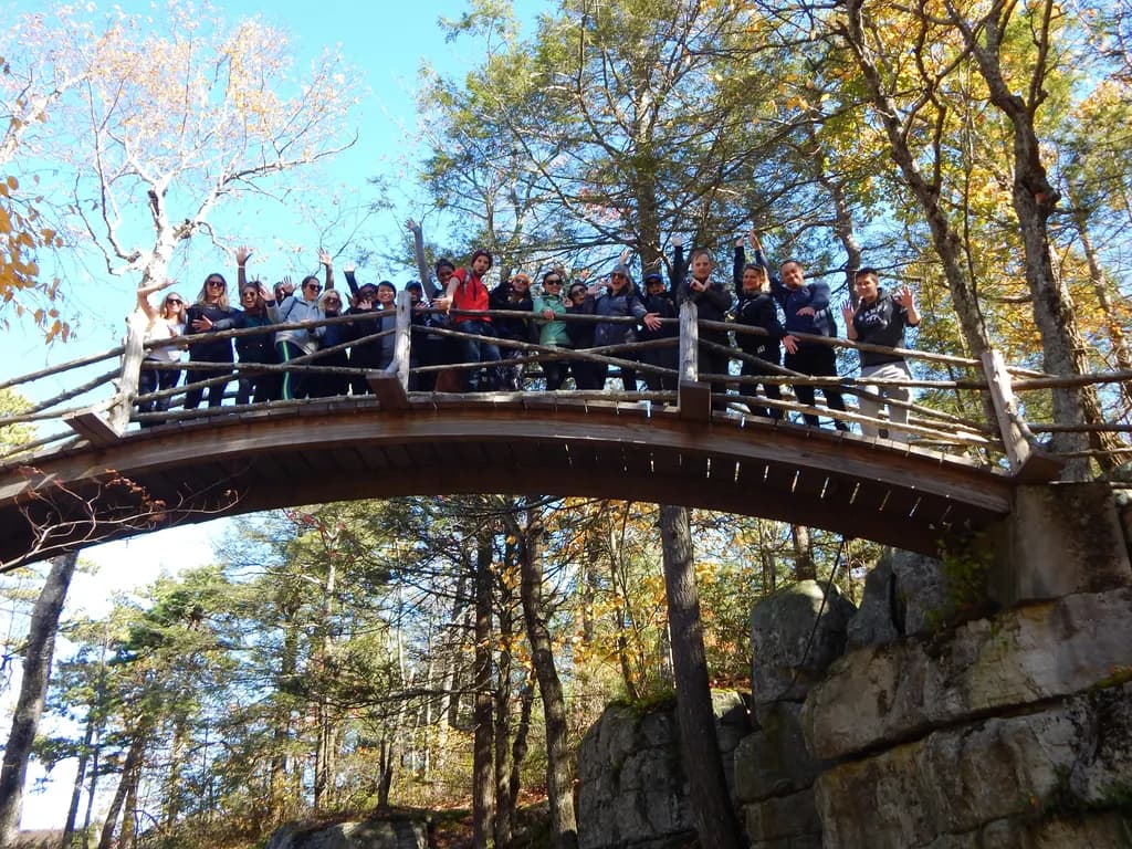 A group of people standing and waving on a wooden bridge in a forested area during autumn.