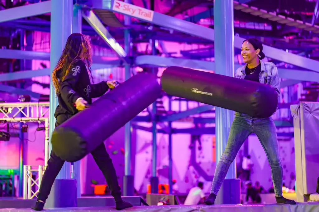 two women using pads to try and knock each other off a beam