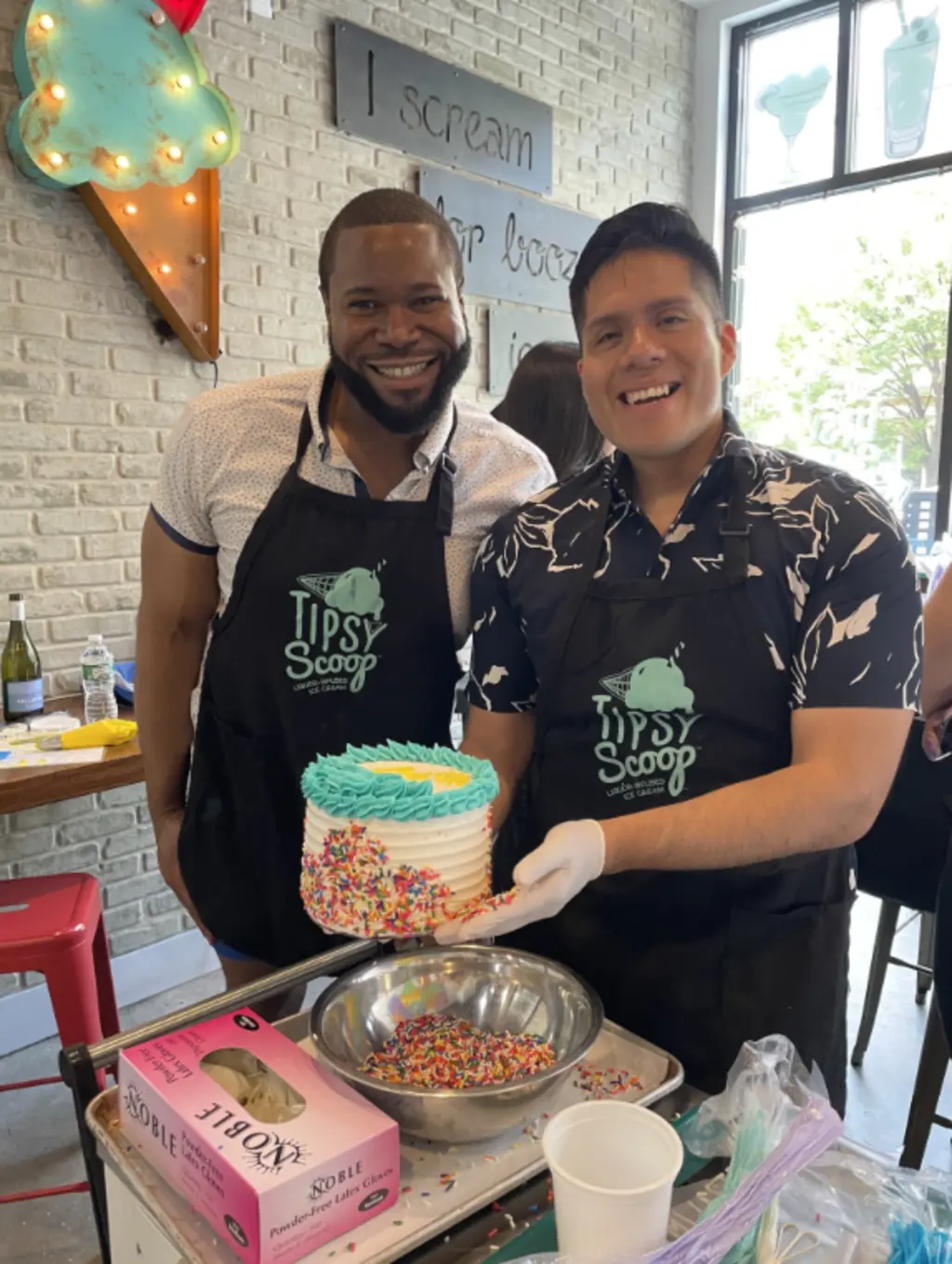 Two men wearing aprons, smiling proudly while holding a decorated cake with rainbow sprinkles during a cake-making class.