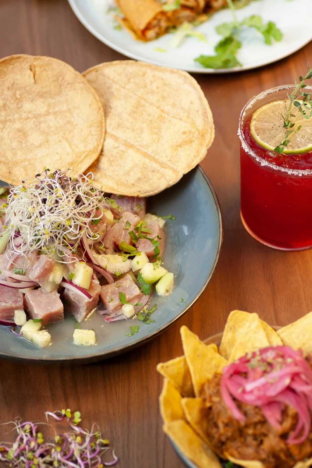 A beautifully plated dish of ceviche with fresh tortillas and garnished with sprouts, accompanied by a vibrant drink, set on a wooden table.