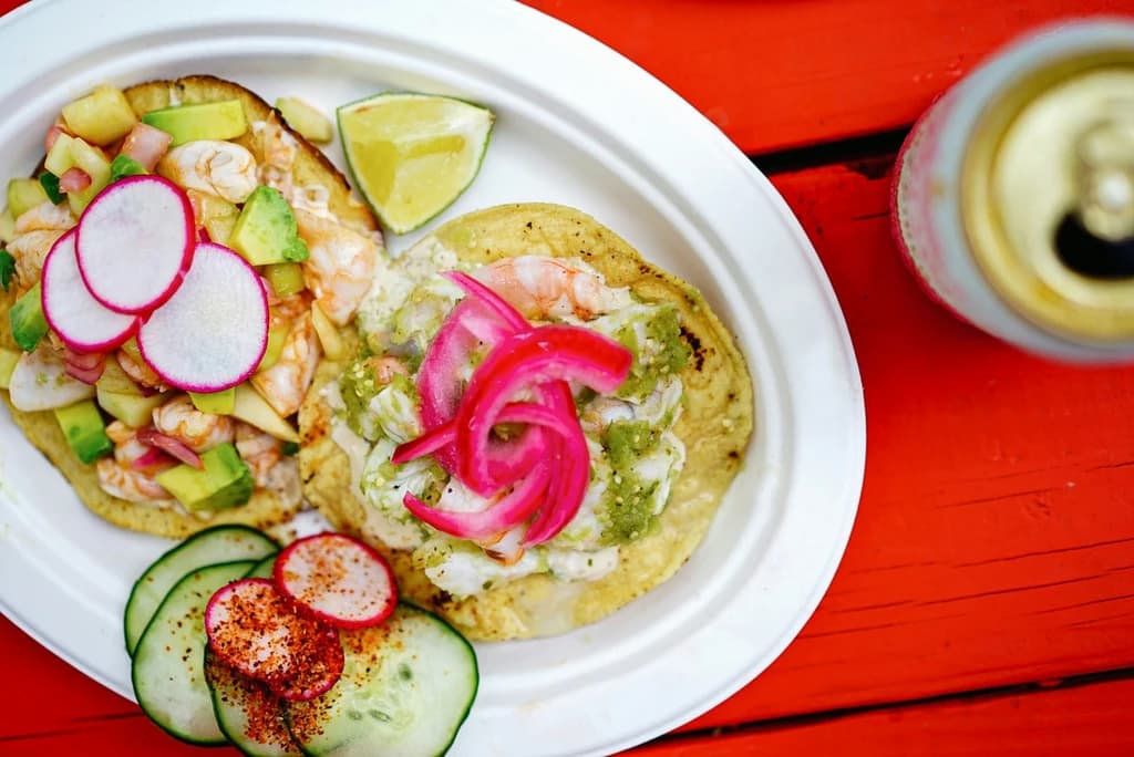 Two colorful tostadas topped with shrimp, avocado, radishes, and pickled onions, served with lime wedges and sliced cucumbers on a bright red table.