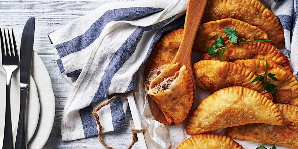 Crispy fried empanadas arranged in a basket with a wooden spatula, garnished with fresh parsley, accompanied by a striped kitchen towel and cutlery on a wooden table.