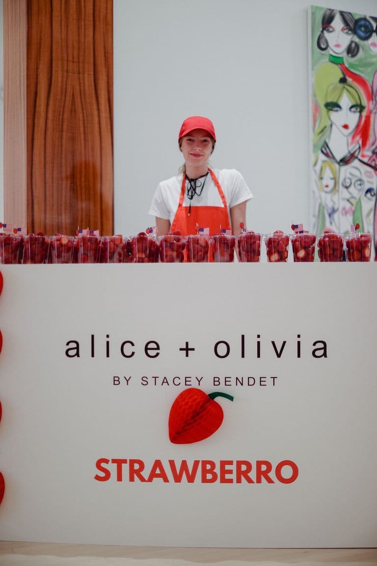 A Strawberryro stand at an Alice + Olivia event, with a smiling attendant serving fresh strawberry cups in a stylish setting.
