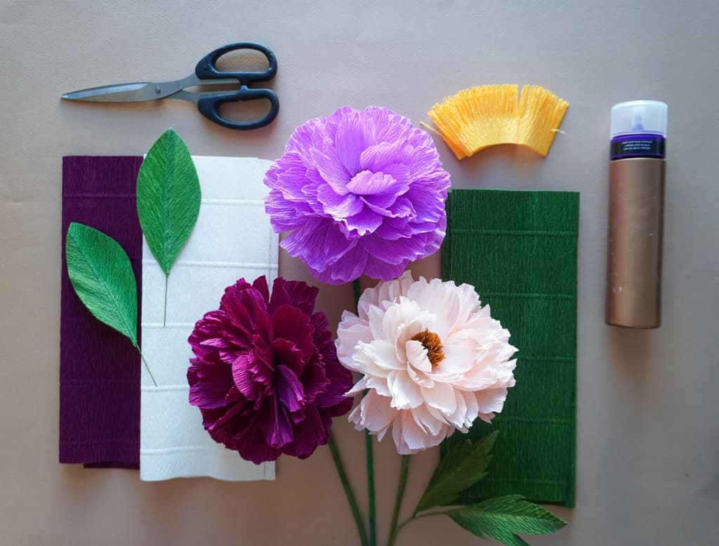 A flat lay of crafting materials including scissors, paper flowers in purple and pink hues, green leaves, and crepe paper, all arranged neatly on a work surface.