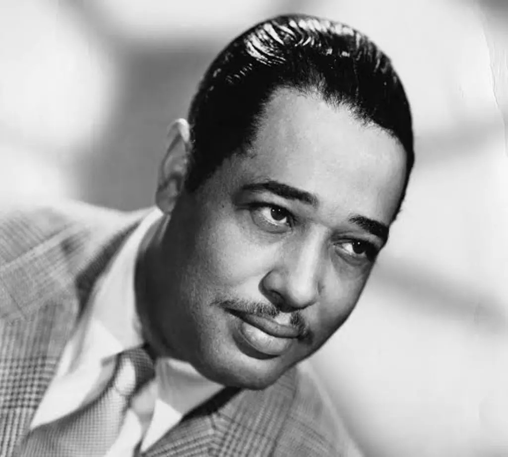 A black-and-white close-up of a man with slicked-back hair and a mustache, smiling slightly while looking to the side.