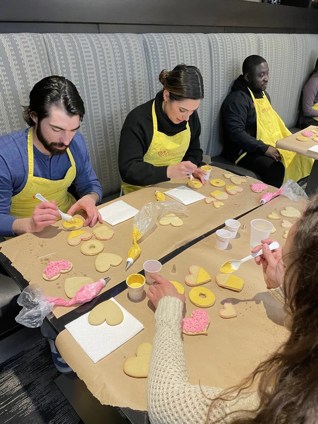 Group of participants decorating heart-shaped cookies with yellow and pink icing at a cookie-making workshop.