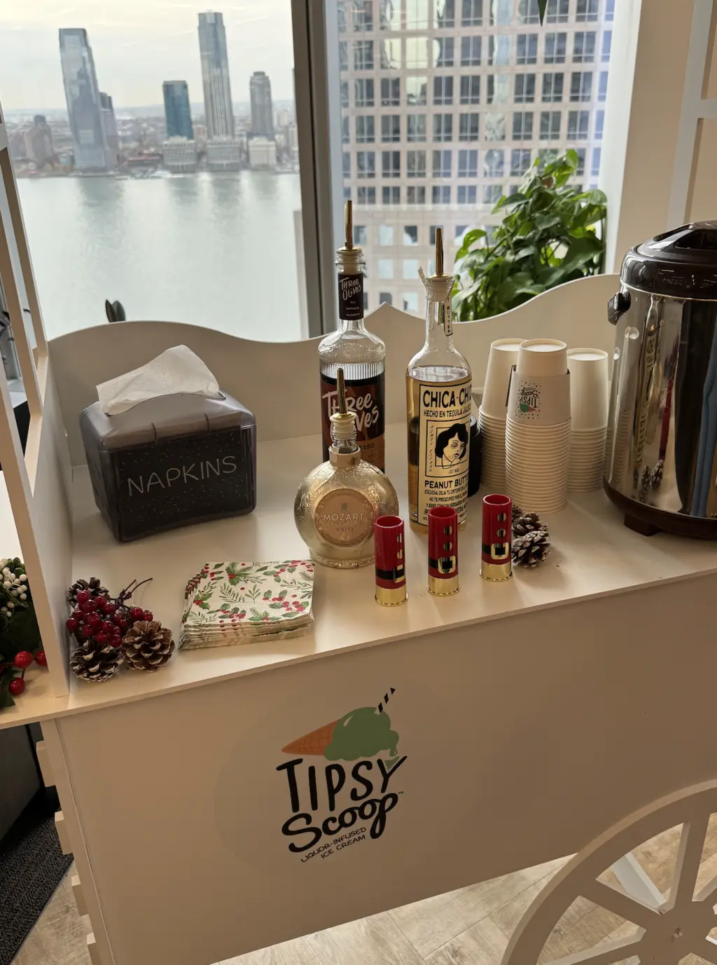 A white Tipsy Scoop cart adorned with holly garlands, cups, and bottles, overlooking a cityscape and waterfront.