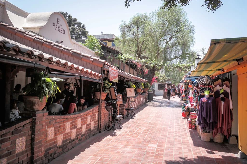 Charming outdoor marketplace with shops and eateries on a sunny day, showcasing traditional Mexican crafts and clothing under shaded awnings and lush greenery.