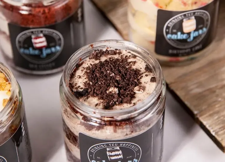 Close-up of assorted cake jars, including cookies & cream, red velvet, and birthday cake, topped with crumbled cookies and colorful sprinkles.