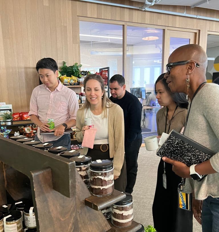 A group of people smiling and browsing a display of layered desserts in jars at an indoor event with snacks and greenery in the background.