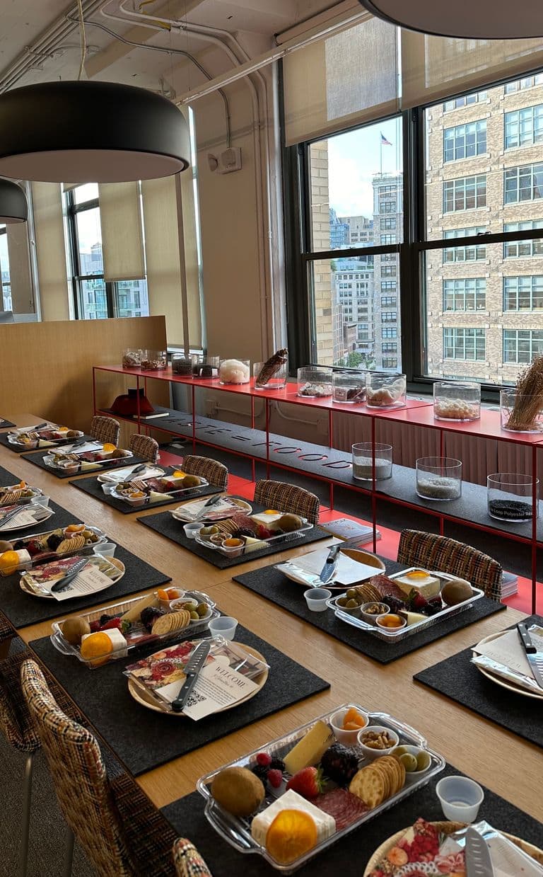 Bright, urban event space with large windows and a long wooden table set with individual charcuterie trays, welcome cards, and knives, surrounded by woven chairs and modern shelving.