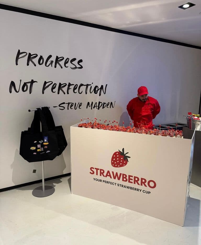 A Strawberryro stand at a Steve Madden event with a bold "Progress Not Perfection" wall quote, offering fresh strawberry cups.