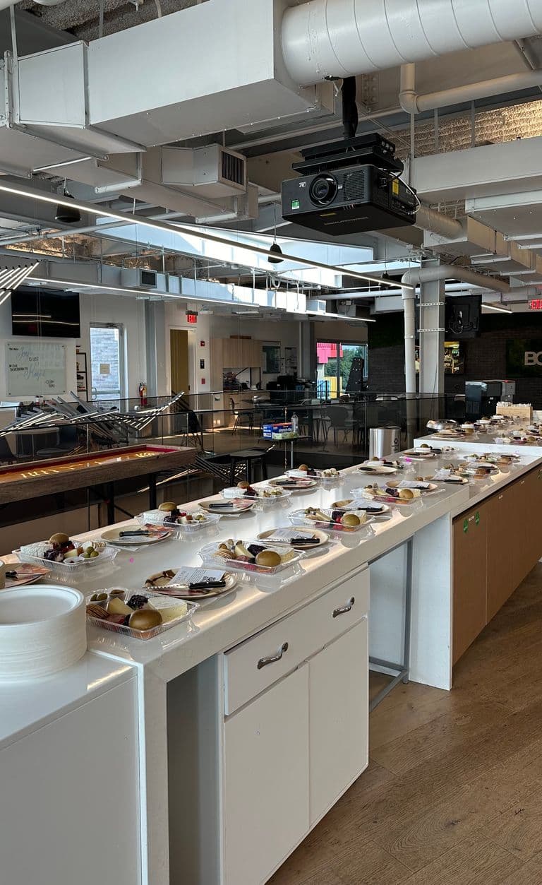 Modern industrial-style event space with exposed ceilings, featuring long counters lined with individual charcuterie trays, plates, and utensils ready for guests.