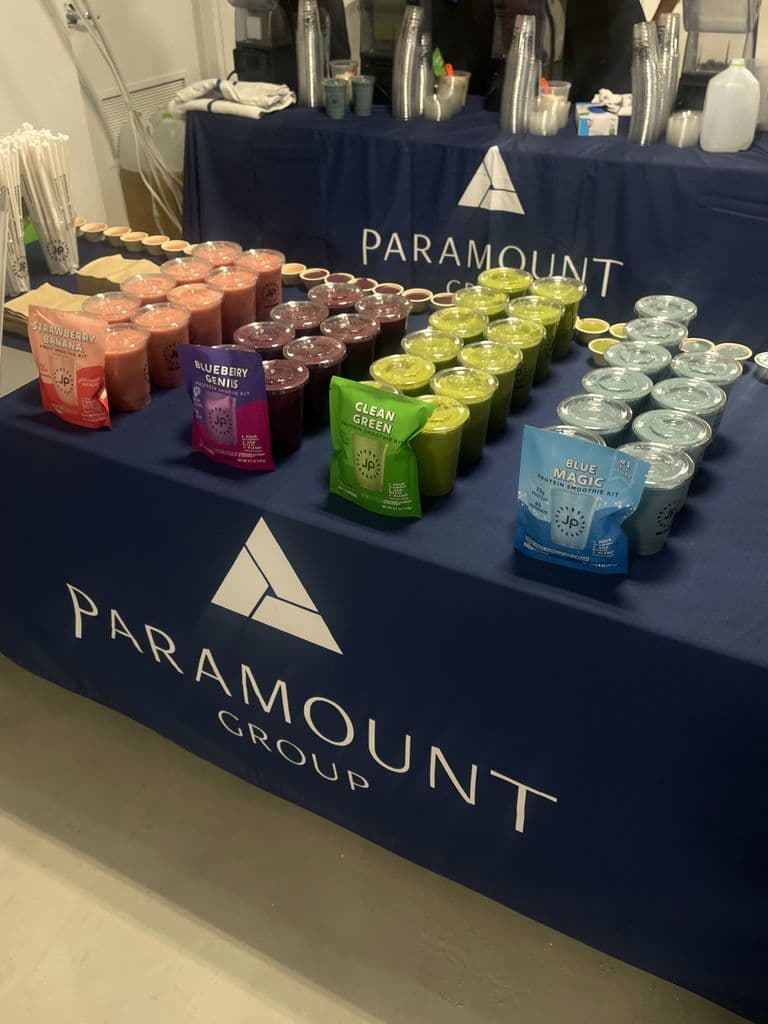 A table with colorful smoothies in red, purple, green, and blue, branded with Paramount Group and JP, set up for an event.