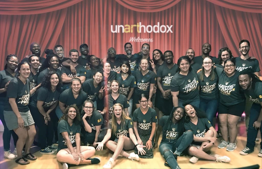 Group of people posing together in matching shirts, standing and sitting in front of a stage with a sign reading 'unarthodox welcomes'.