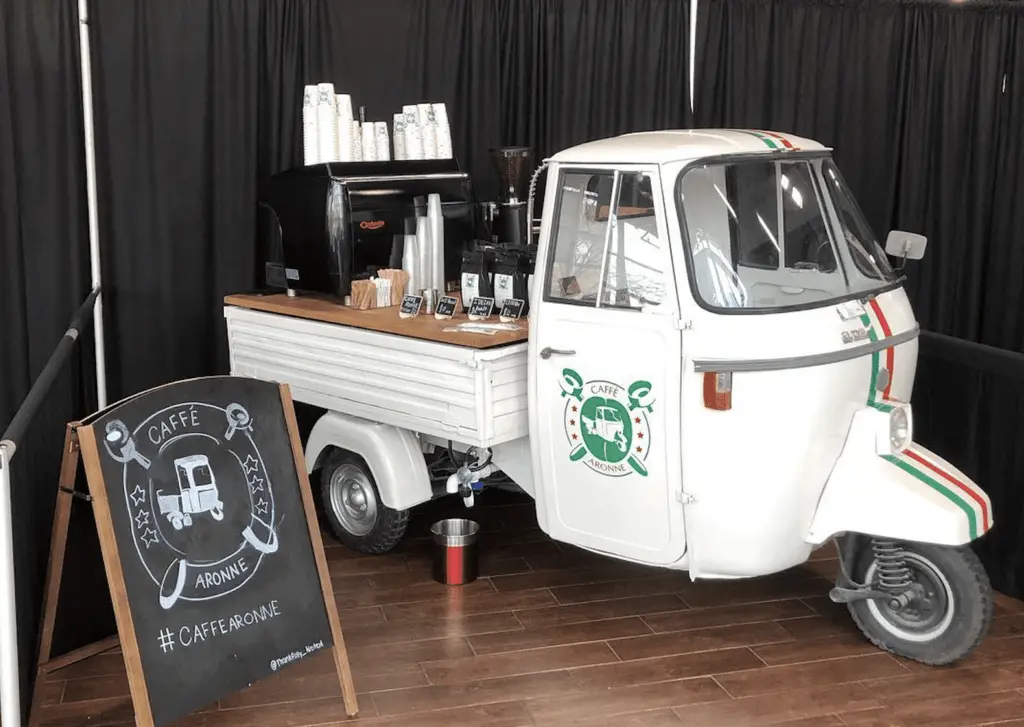 An Italian-style coffee cart with a vintage look, featuring a white three-wheeled truck serving coffee. A chalkboard sign reads Caffè Aronne.