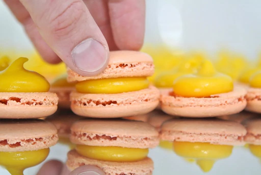 A close-up of a hand assembling peach-colored macarons with bright yellow filling, reflected on a glossy surface.