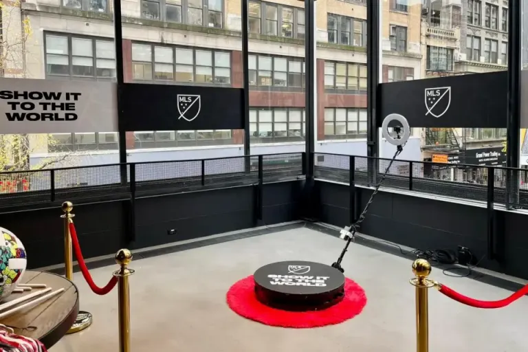 A 360-degree photo booth setup branded with MLS, featuring a rotating camera arm, a ring light, and red velvet ropes in an urban event space.