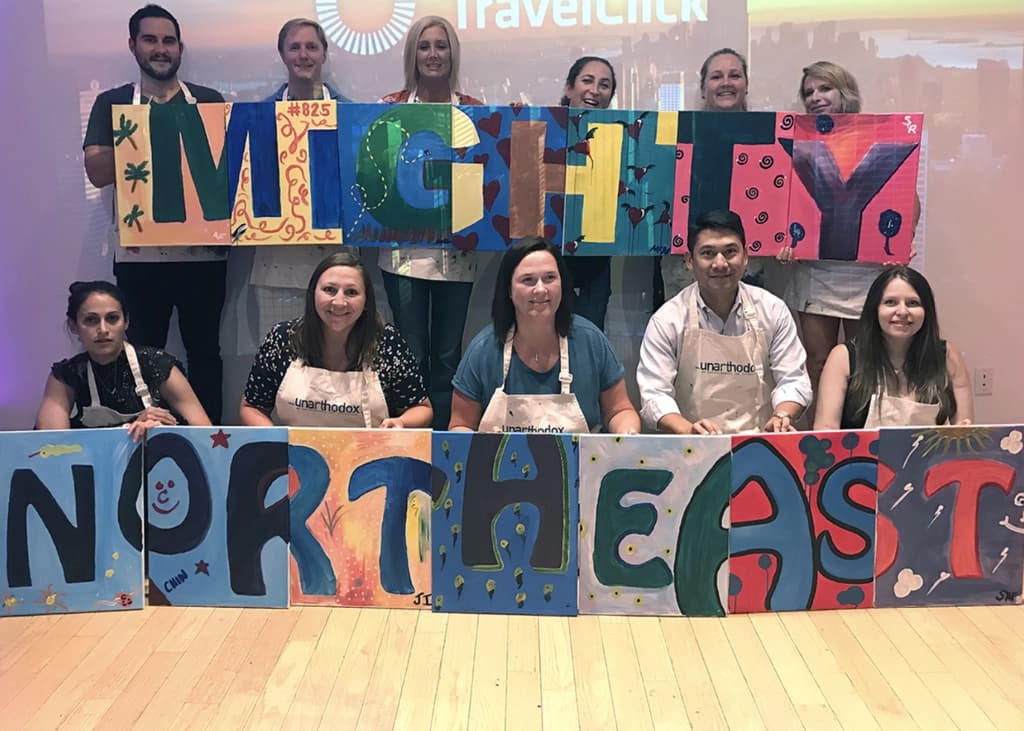 Participants standing and sitting around a completed mural, each holding different sections of their colorful group painting.