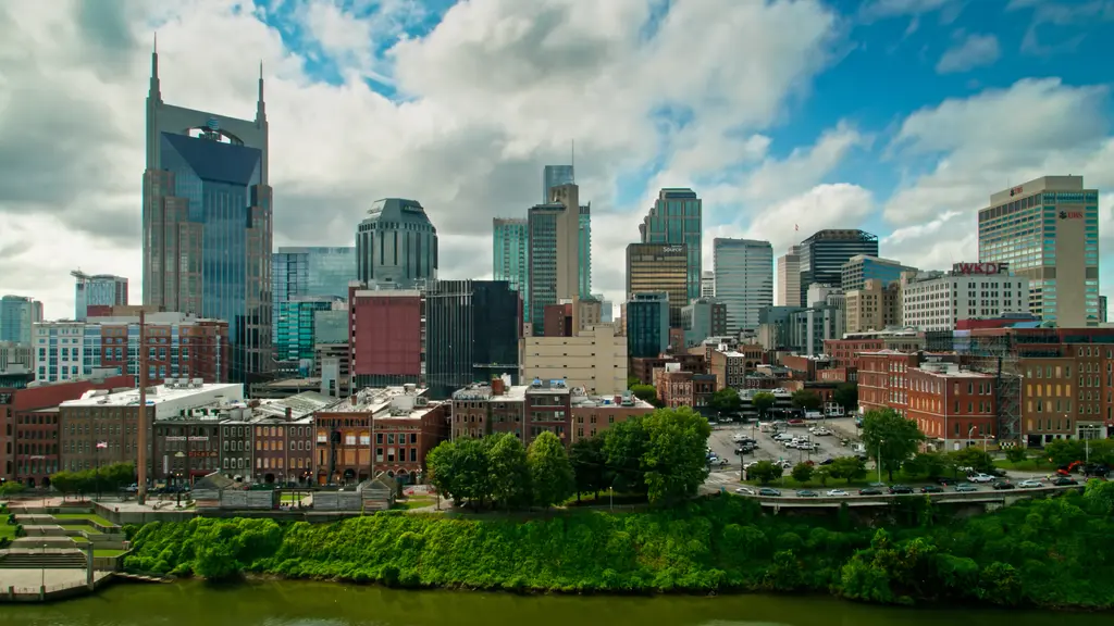 View of the Nashville city skyline on a cloudy day, with tall buildings, historic architecture, and green trees lining the riverbank.