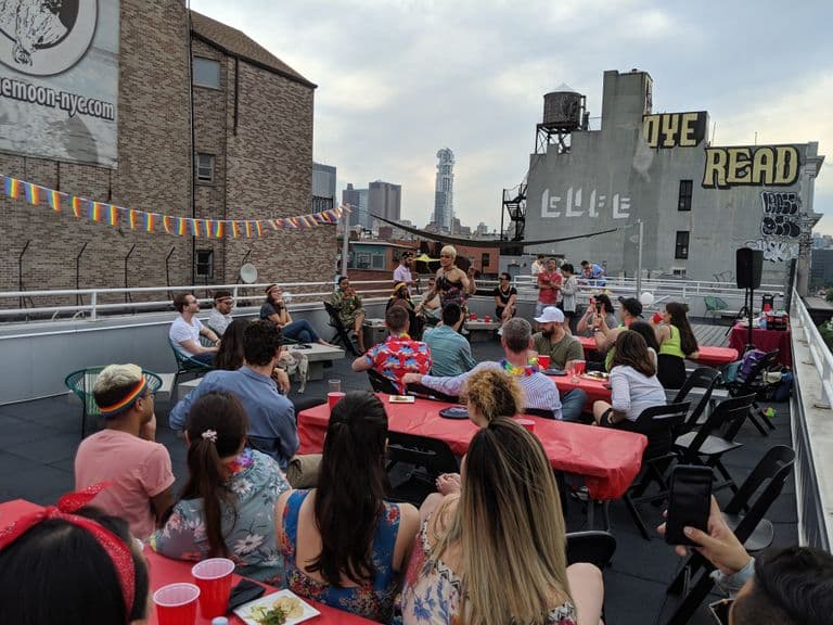 "A rooftop gathering in New York City where a performer with short blonde hair is entertaining a seated audience. Attendees sit at tables with red tablecloths, many wearing colorful summer attire and pride accessories. Rainbow flag bunting is strung across the space. The building on the right has 'DYE' and 'READ' visible in yellow lettering on its gray wall. In the background, the NYC skyline is visible with a distinctive tall residential tower. The event appears to be a pride celebration or drag performance taking place at dusk."Retry