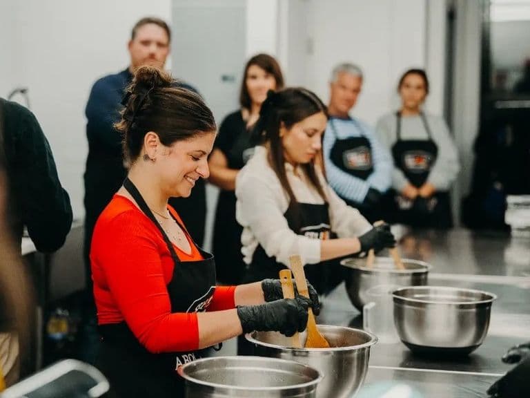 People in aprons and gloves smile while mixing ingredients during a hands-on cooking class.