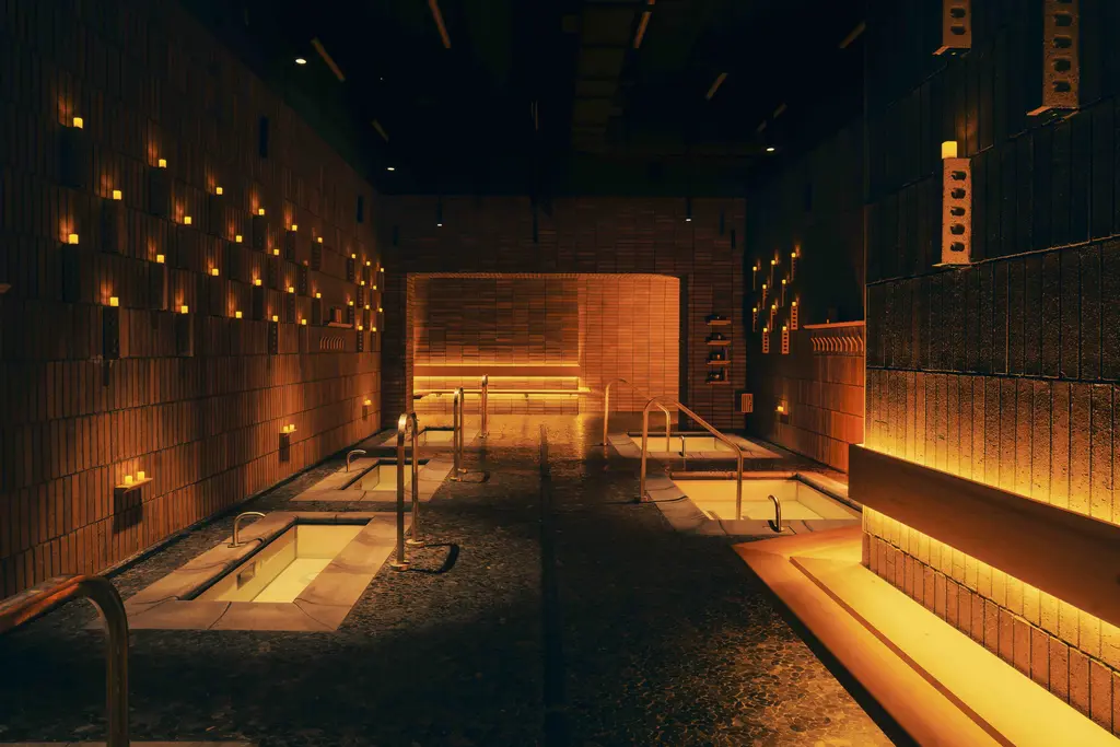 An atmospheric spa room with a long pool surrounded by warm, ambient lighting, brick walls, and candles, creating a serene and relaxing environment.
