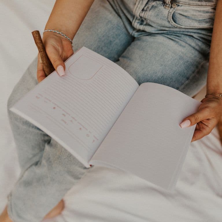 Person holding an open journal and pen, seated cross-legged in light denim jeans.