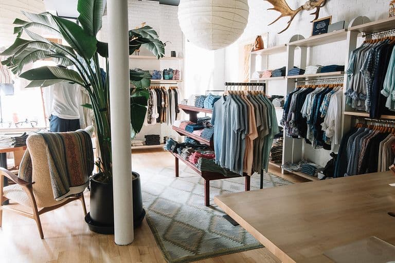 Bright and airy Faherty store interior with natural light, a large potted plant, cozy seating, and neatly folded casual shirts and apparel on display racks.