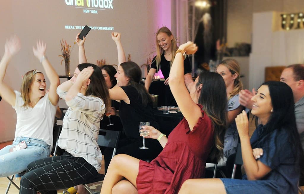 A group of women cheering and raising their hands in excitement while seated, enjoying a team-building event with drinks and laughter.