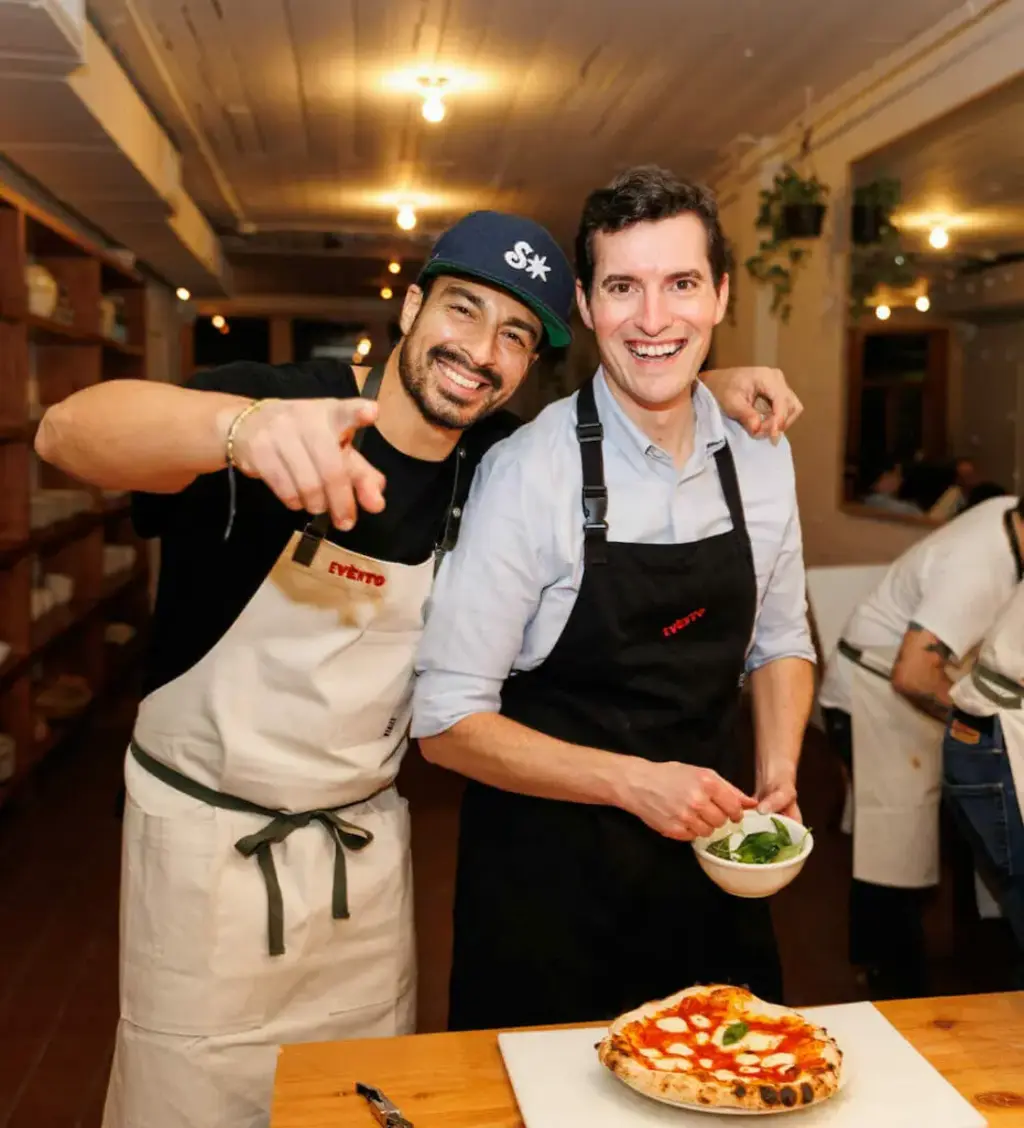 a person standing with an instructor, smiling and putting toppings on their pizza