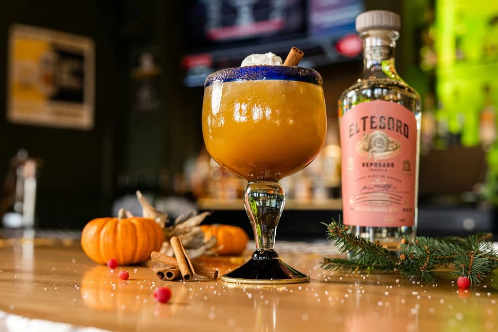 A margarita glass with a pumpkin-colored drink, whipped cream topping, surrounded by pumpkins and a bottle of El Tesoro tequila.