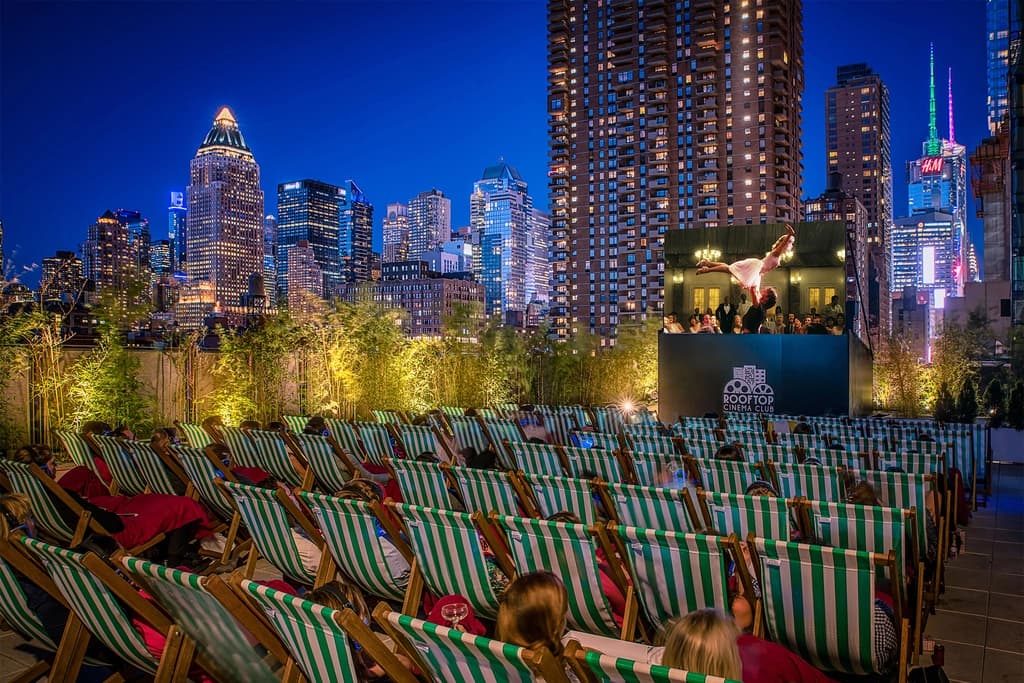 A rooftop cinema with people in striped lounge chairs watching a movie against a stunning backdrop of city skyscrapers under a clear night sky.