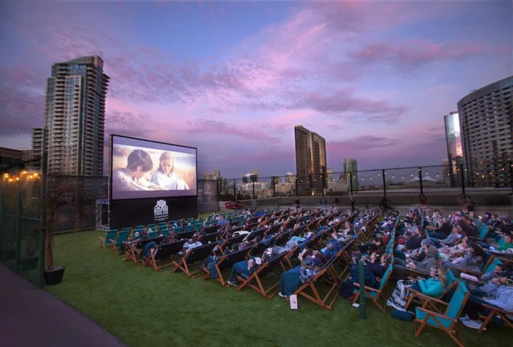 An outdoor rooftop cinema with people in lounge chairs watching a movie at sunset, with city skyscrapers in the background under a colorful sky.
