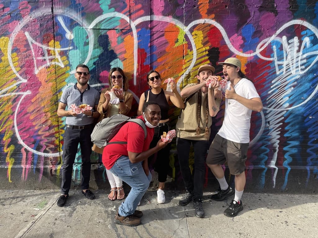 A group of six people posing in front of a vibrant graffiti wall, smiling and holding sandwiches, enjoying a food tour.