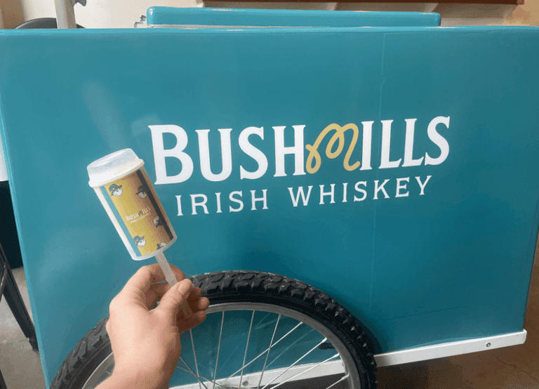 A hand holds a Bushmills-branded push pop in front of a teal Bushmills Irish Whiskey cart with a large logo and a visible bicycle wheel.