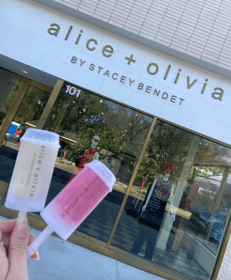 Two Alice + Olivia branded popsicles held in front of the store’s entrance, with mannequins visible through the glass doors.