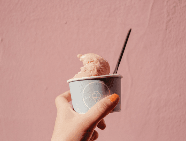 Hand holding a cup of pink ice cream with a black spoon in front of a pastel pink wall.