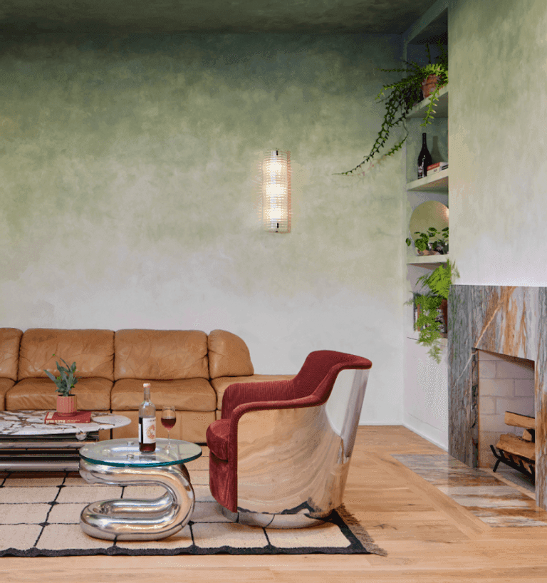Cozy lounge with a tan leather couch, red chrome chair, sculptural table, fireplace, and plants on built-in shelves.