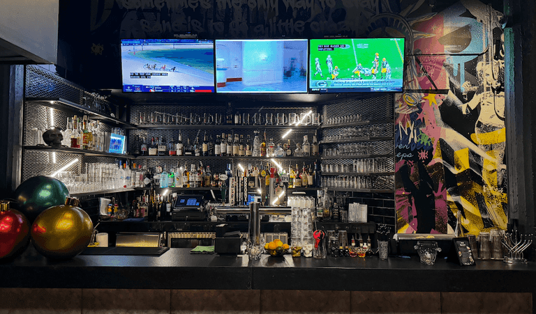 A vibrant bar setup with colorful graffiti wall, TVs showing sports, and shelves full of bottles and glassware.
