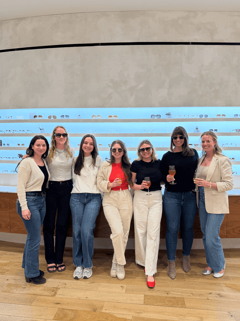 Seven women stand smiling in front of a stylish eyewear display, each holding a drink and wearing sunglasses in a modern showroom.