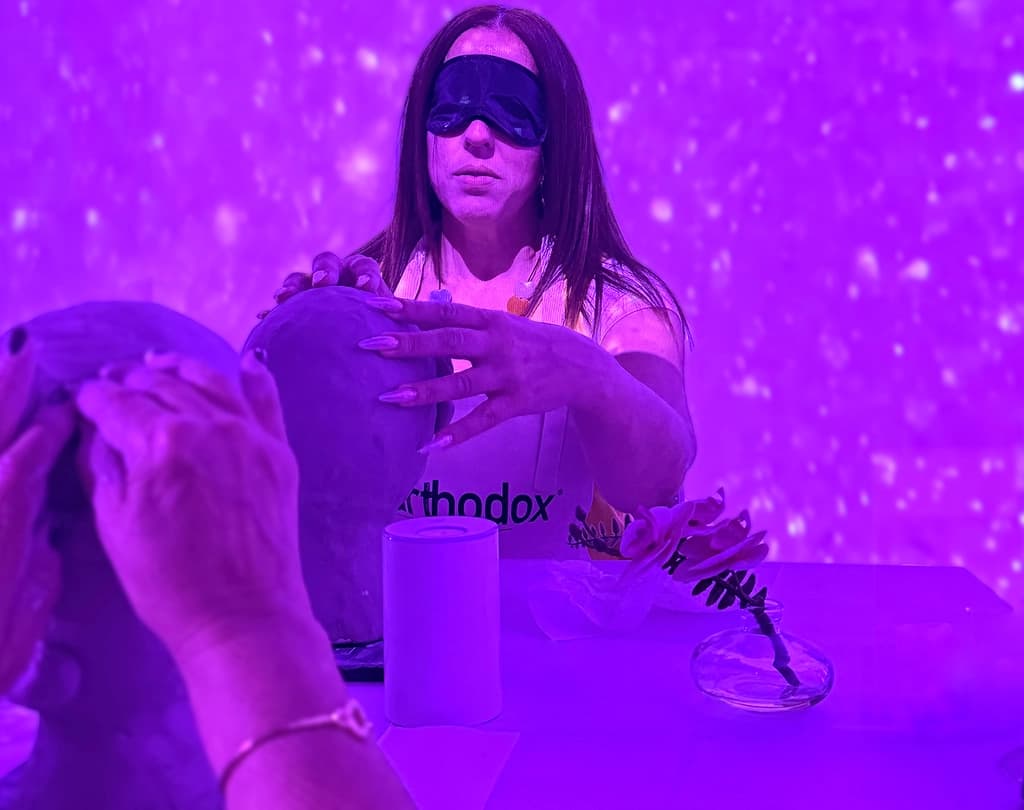 A blindfolded woman sculpting a clay head in a neon-lit room, surrounded by soft, glowing visuals, fully immersed in the creative process.
