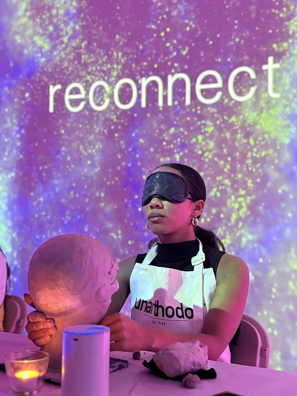 A woman wearing a blindfold, sculpting a clay head with intense focus under purple lighting in an immersive activity.