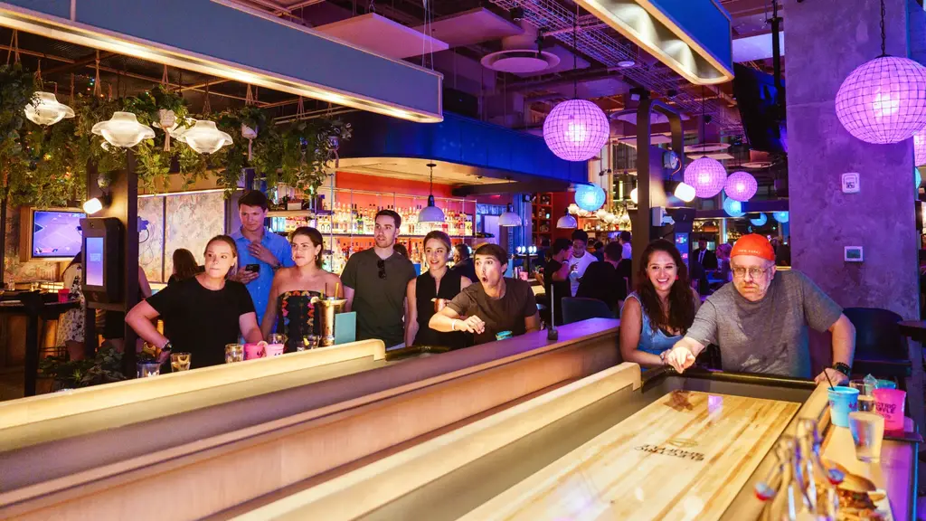 Group of people enjoying a game of shuffleboard in a vibrant bar setting with colorful lighting and decor.