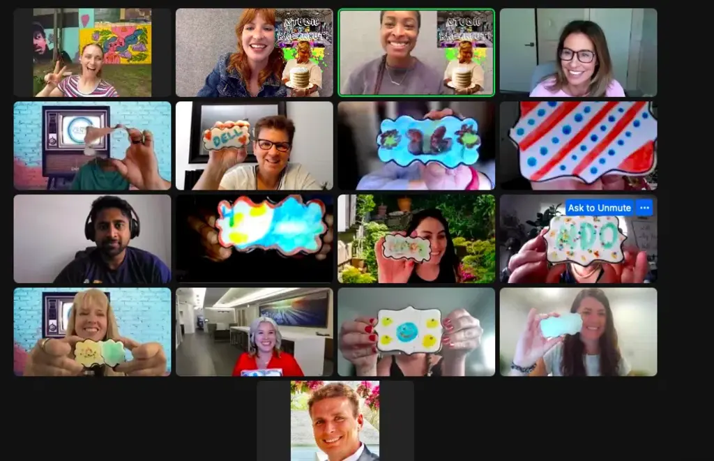 A virtual group of 16 people smiling, holding up their creatively decorated cookies in a Zoom meeting, showcasing their designs.