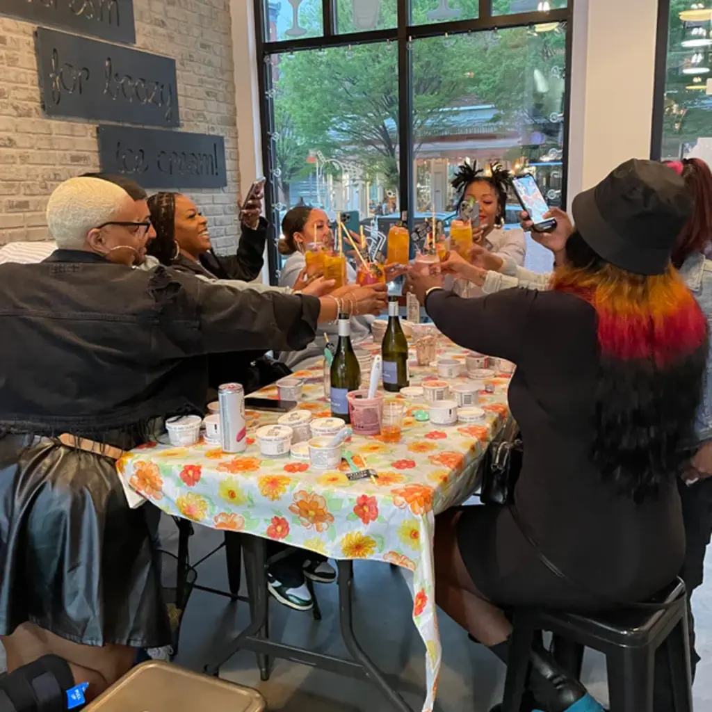 A group of friends cheers with drinks around a table decorated with ice cream cups and bottles, enjoying a sundae-making event.